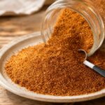 Reddish-brown sweet and spicy dry rub beside a clear jar and metal measuring spoon