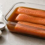Raw salmon fillet pieces soaking in brown sugar vinegar brine with salt and ground allspice beside a glass container