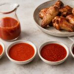 Four bowls of red chicken wing sauce arranged from mild to nuclear beside a glass pitcher of the tomato hot sauce base