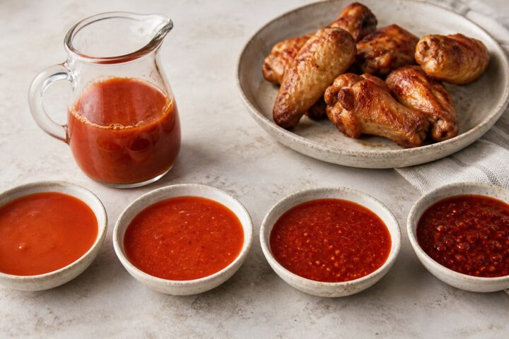 Four bowls of red chicken wing sauce arranged from mild to nuclear beside a glass pitcher of the tomato hot sauce base