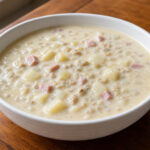 Creamy clam chowder with diced potatoes and ham in a white ceramic bowl on a wooden table