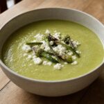 Silky green garlic scape soup topped with crumbled feta and grilled scape flower tops in a ceramic bowl