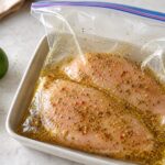 Two chicken breasts marinating with lime zest, oregano, coriander, and red pepper in a clear food bag