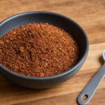 Reddish-brown smoky steak dry rub with oregano flecks in a dark ceramic bowl beside a measuring spoon