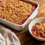 Golden oat crumble over pink-red diced rhubarb in a square baking dish with one warm serving alongside