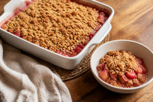 Golden oat crumble over pink-red diced rhubarb in a square baking dish with one warm serving alongside
