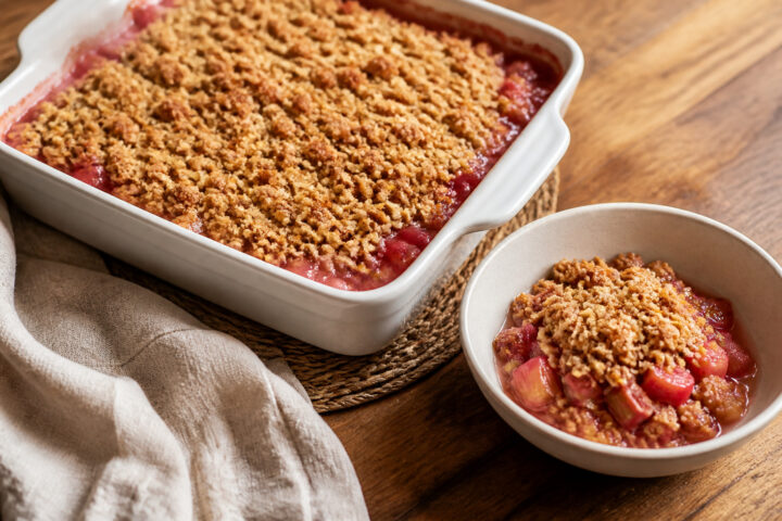 Golden oat crumble over pink-red diced rhubarb in a square baking dish with one warm serving alongside