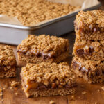 Golden oatmeal carmelita bars with visible caramel, semisweet chocolate, and chopped pecan layers on a warm table