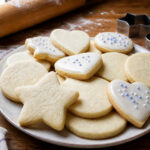 Thick pale cutout sugar cookies in simple shapes, some plain and some lightly iced, beside a rolling pin and cutters