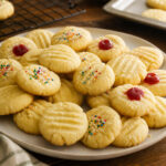 Small pale whipped shortbread cookies with fork marks, restrained nonpareils, and a few candied cherry halves