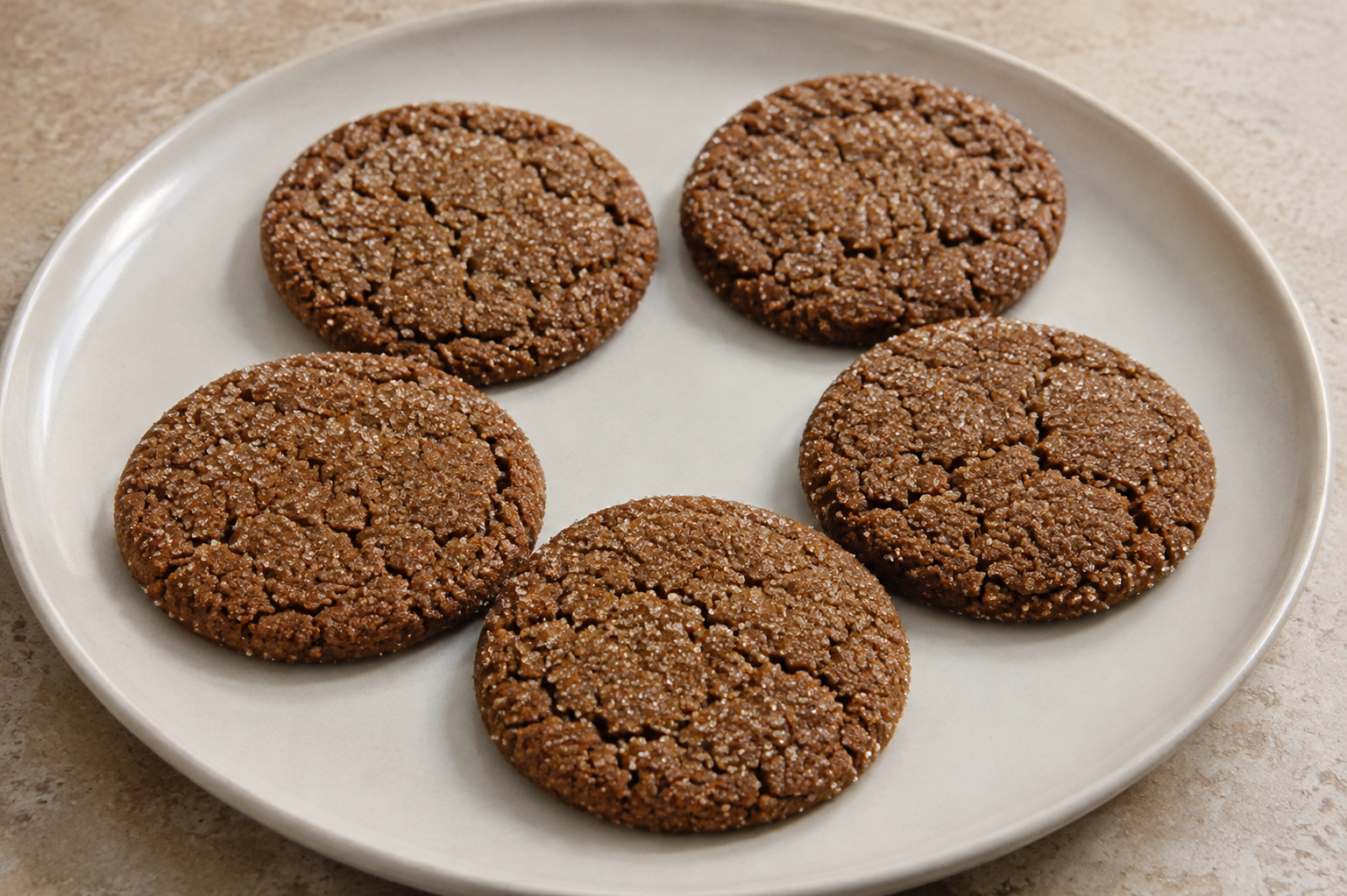 Five brown molasses ginger cookies with cracked tops and sparkling granulated sugar on a white plate.
